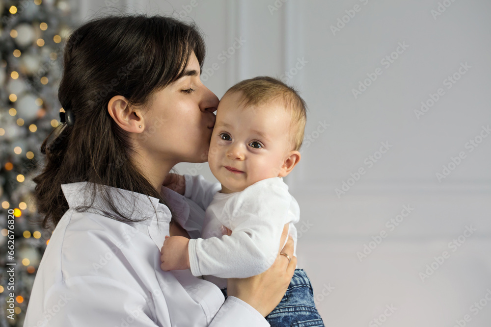 A young mother kisses her little child while standing near a Christmas tree A young mother kisses her little child while standing near a Christmas tree