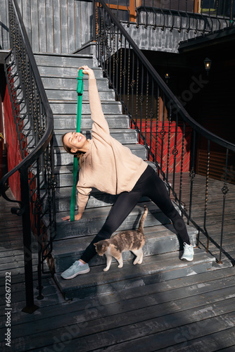 Satisfied young woman with her cat plays sports on the street, performs the Trikonasana exercise, triangle pose with a fitness belt, practices yoga in sportswear standing on the staircase thresholds