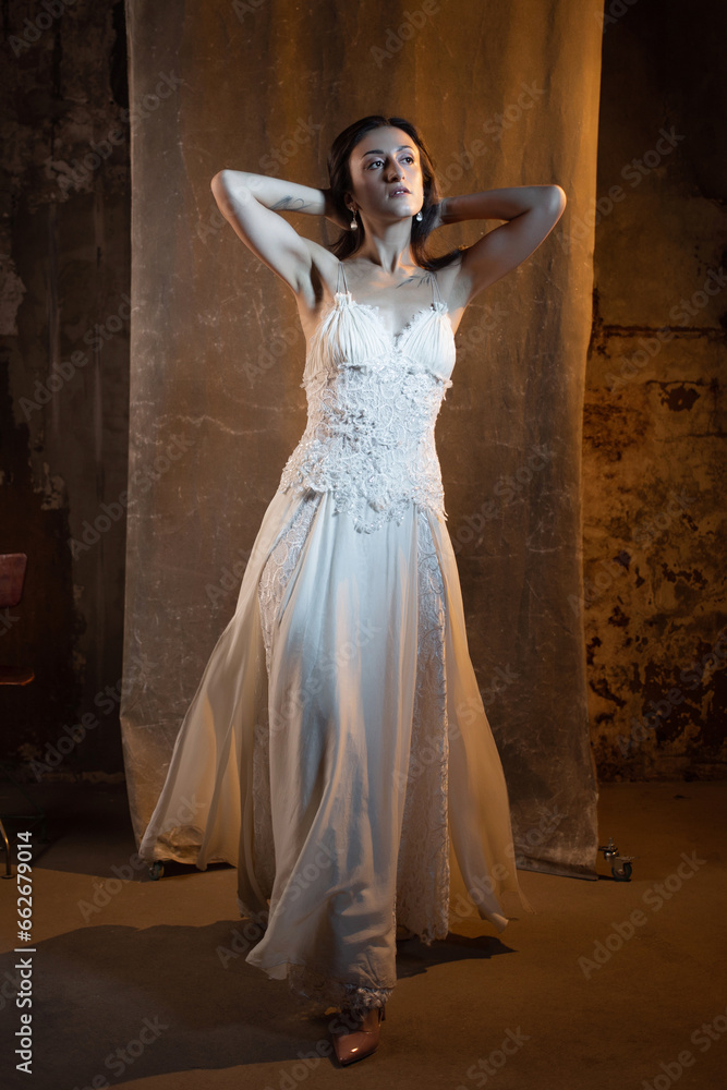 Elegant and beautiful brunette in a chic evening dress or wedding dress, white dress with a long flowing skirt to the floor. photo in dark colors on a textured wall, warm light Elegant and beautiful brunette in a chic evening dress or wedding dress, white dress with a long flowing skirt to the floor. photo in dark colors on a textured wall, warm light