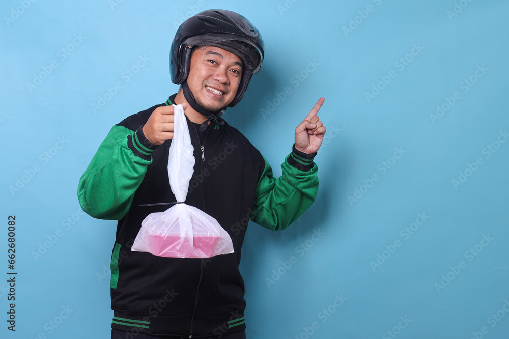 Asian online taxi driver wearing jacket and helmet holding food box in plastic bag while pointing at empty space for text or ads Asian online taxi driver wearing jacket and helmet holding food box in plastic bag while pointing at empty space for text or ads