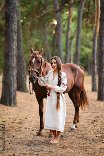 Beautiful extra long braided hair girl in Ukrainian traditional dress posing with horse in forest . Portrait of young attractive stylish woman on colorful warm background. © Ольга Бойко