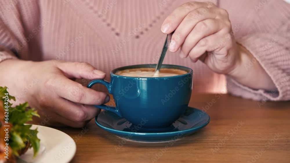 A Hand is Mixing the Milk Foam of Latte. Person Stirring Cappuccino with Steel Spoon. Hot Coffee in a Dark Blue Ceramic Cup on a Wooden Table. Modern Coffee shop, Cafeteria. Thick Froth. Close-up View A Hand is Mixing the Milk Foam of Latte. Person Stirring Cappuccino with Steel Spoon. Hot Coffee in a Dark Blue Ceramic Cup on a Wooden Table. Modern Coffee shop, Cafeteria. Thick Froth. Close-up View