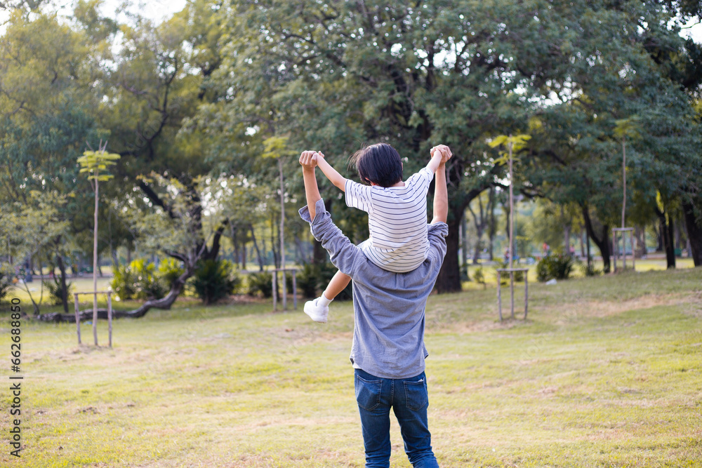 Asian playful boy sitting on father shoulder playing at park in summer. Dad raise kid hand up in the air and running. happy family lifestyle insurance