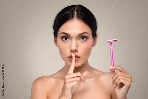 Serious calm millennial caucasian woman shows razor, puts finger to lips, makes shhh sign photo