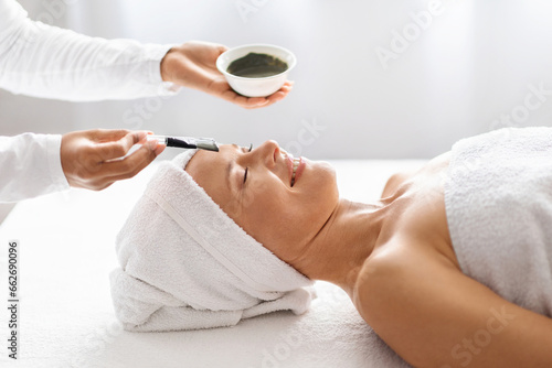 Skilled beautician applying moisturizing clay mask to middle aged woman at spa
