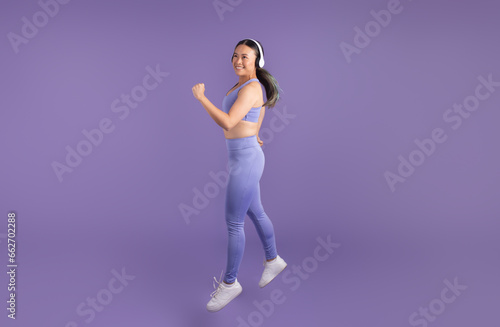 Asian woman in headphones jumping over purple studio backdrop, full length