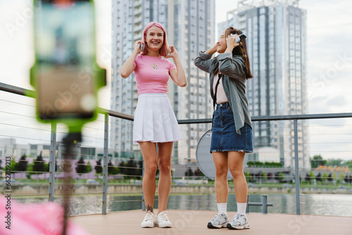 Two smiling girls making video with smartphone while standing on city waterfront