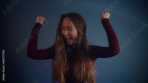 Portrait of a young beautiful smiling happy jubilant pretty caucasian girl with raised hands, long brown hair in a burgundy sweater on a blue background. Emotions of triumph, joy and jubilation