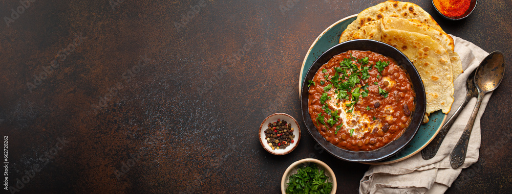 Traditional Indian Punjabi dish Dal makhani with lentils and beans in black bowl served with naan flat bread, fresh cilantro and two spoons on brown concrete rustic table top view. Space for text. - obrazy, fototapety, plakaty 