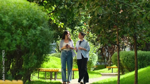 Smiling female friends talking while walking outdoors. Two young women having conversation in the park, pretty girls chatting and gossiping. Business women share ideas during work break