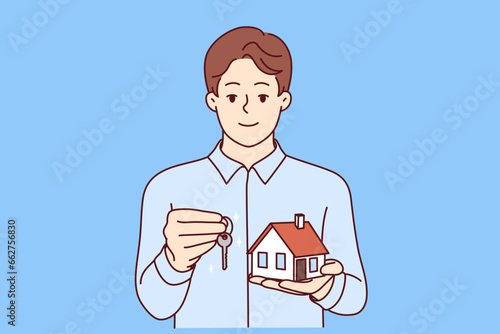 Man real estate broker holds keys and model house offering to take out mortgage on favorable terms. Real estate agent guy looks at screen and smiles, recommending to provide consultation services