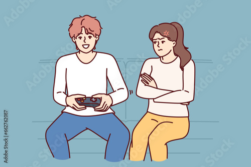 Woman is angry at husband who plays video game and does not want to do housework. Disgruntled girl sits on couch near boyfriend who is keen on video game, persuading him to go for walk
