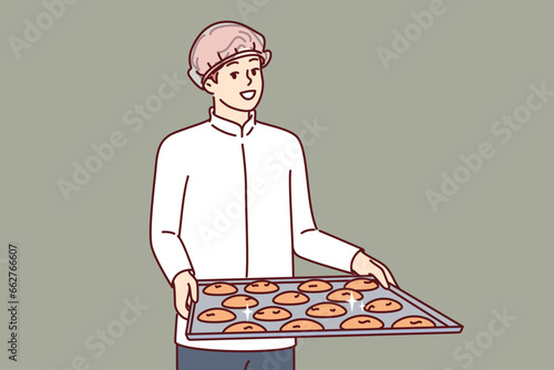 Man pastry chef is holding tray of cookies, dressed in white chef shirt and disposable hair cap. Guy confectioner prepares oatmeal cookies from natural ingredients, for sale in own bakery