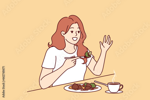 Woman is having lunch in cafe sitting at table and raising hand up to call waiter. Cafe visitor girl eats steamed vegetables to avoid passing fatty and caloric food causing weight gain