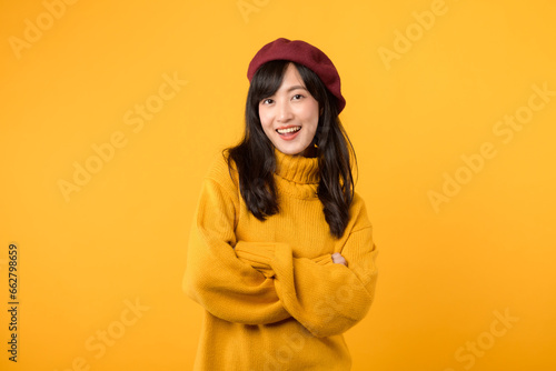 Positive young Asian woman in her 30s, with arms crossed on her chest, exuding confidence in a yellow sweater and red beret against a yellow background.