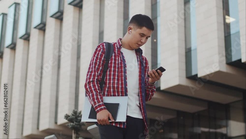 Young asian man with a backpack and a laptop in his hands walking alongside the building, reading correspondence with a client on a smartphone and satisfiedly smiling. Concept of modern remote work photo