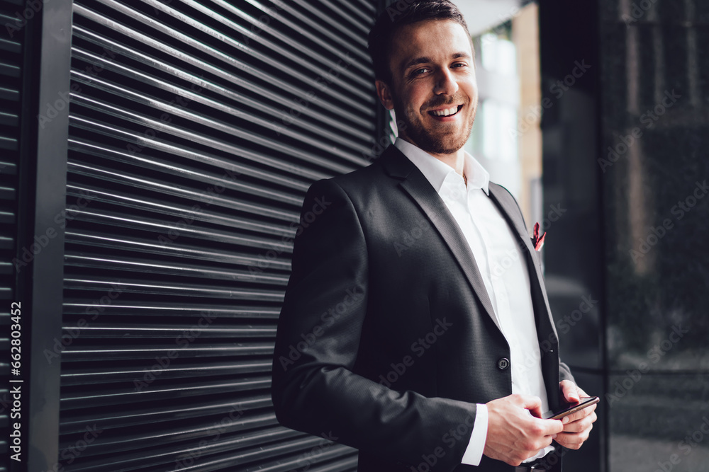 Fototapeta premium Pleasant man in classical suit with smartphone looking at camera with smile in street in downtown