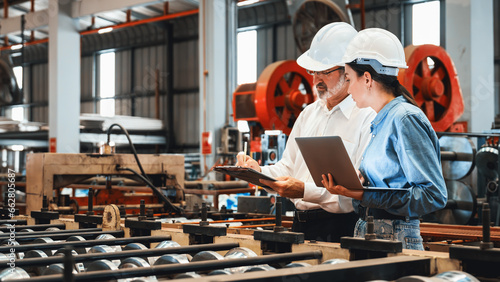 Factory engineer manager with assistant using laptop to conduct inspection of steel industrial machine, exemplifying leadership as machinery engineering inspection supervisor in metalwork manufacture.