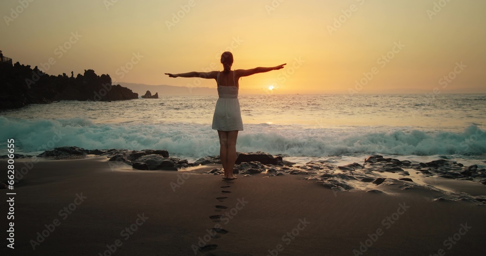Silhouette woman at sunset raising arms in air. Ocean waves crashing on sandy beach and spraying. Concept emotions, joy, happiness.
