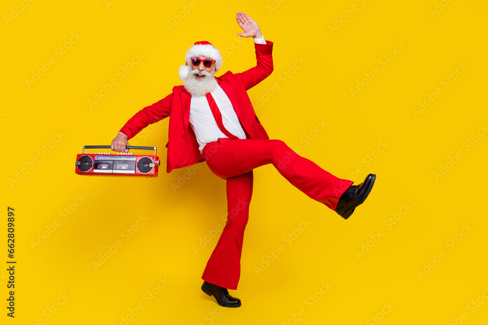 Full length photo of good mood claus wear red tux costume listening boom box walking empty space isolated yellow color background Full length photo of good mood claus wear red tux costume listening boom box walking empty space isolated yellow color background