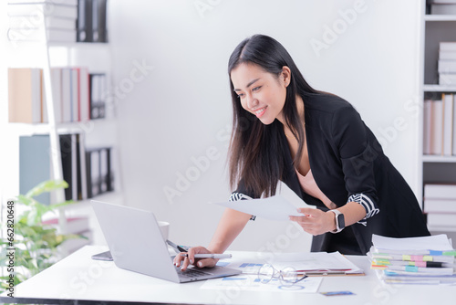 Sharing good business news. Attractive young businesswoman talking on the mobile phone and smiling while sitting at her working place in office and looking at laptop PC. © Tj