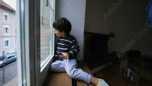 Child's daydream sitting by window at apartment breathing into glass creating vapor. Bored little boy's curiosity observing from second floor building
