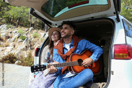 Happy hipster couple sitting in the trunk of their car and singing a songs to the acoustic guitar. Close up, copy space, background.
