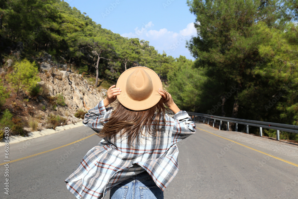 Back view of a free spirited young woman walking down the empty winding road and holding on to her hat. Close up, copy space, background.