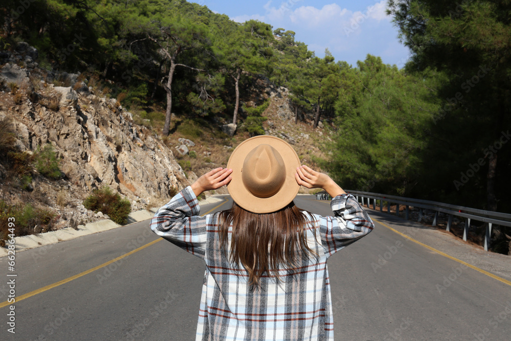 Back view of a free spirited young woman walking down the empty winding road and holding on to her hat. Close up, copy space, background.