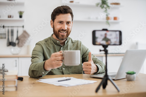 Happy attractive streamer sitting at table near modern laptop and recording video content on smartphone fixed on tripod showing thumb up