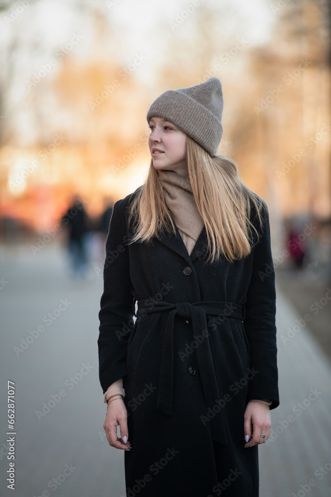 Portrait of a young beautiful blonde girl in a knitted hat and dark coat in an urban environment. Portrait of a young beautiful blonde girl in a knitted hat and dark coat in an urban environment.