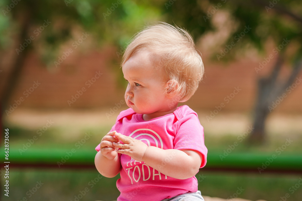 Cute Caucasian blonde toddler girl in pink sweatshirt on the playground in summer Cute Caucasian blonde toddler girl in pink sweatshirt on the playground in summer