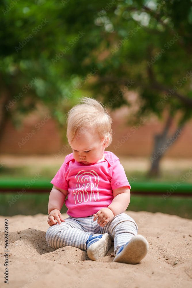 Cute Caucasian blonde toddler girl in pink sweatshirt on the playground in summer Cute Caucasian blonde toddler girl in pink sweatshirt on the playground in summer