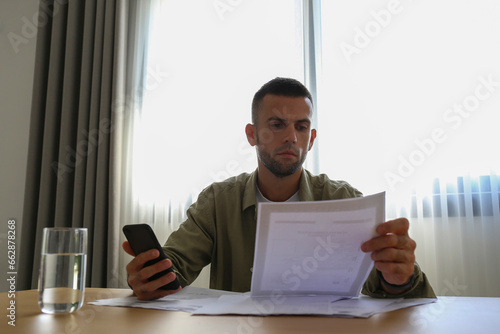 Unamused bearded man looking through his monthly statement and bills at home. A guy sitting by the desk and doing paperwork with the budget app on the phone. Close up, copy space, interior background.