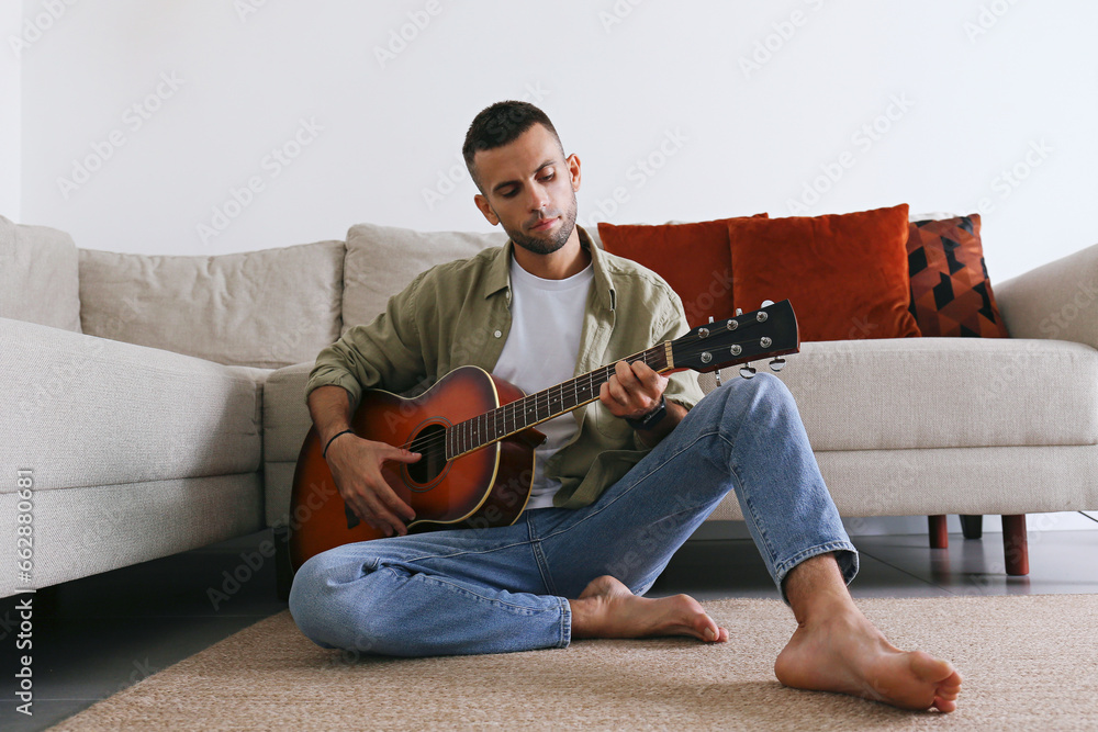 Obraz premium Young man sitting on the floor and playing dreadnought parlor acoustic guitar at home. Guitarist with sunburst colored instrument. Background, copy space, close up