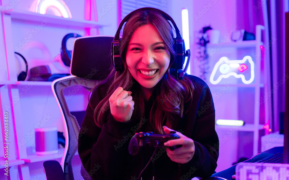 Beautiful happy young female game online streamer or caster smiling with fun, succeed for winner, using computer to play, sitting in room with decorated neon light. Looking at camera with copy space. Beautiful happy young female game online streamer or caster smiling with fun, succeed for winner, using computer to play, sitting in room with decorated neon light. Looking at camera with copy space.