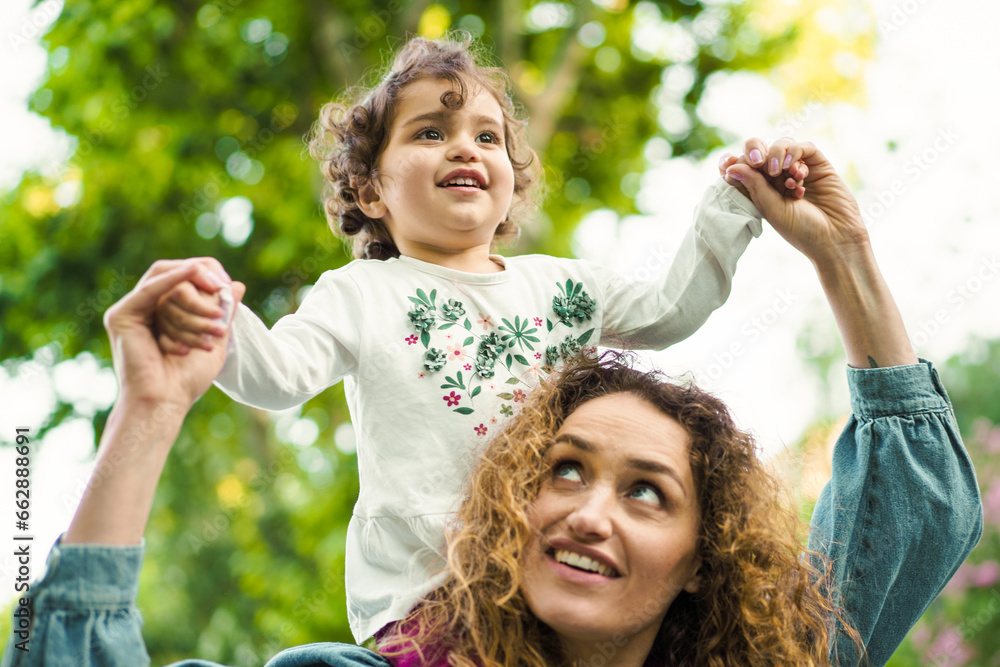 Obraz premium Joyful Moments in Nature: A cheerful young child with curly hair enjoys a playful moment, being lifted up by a loving woman, surrounded by the beauty of lush greenery.