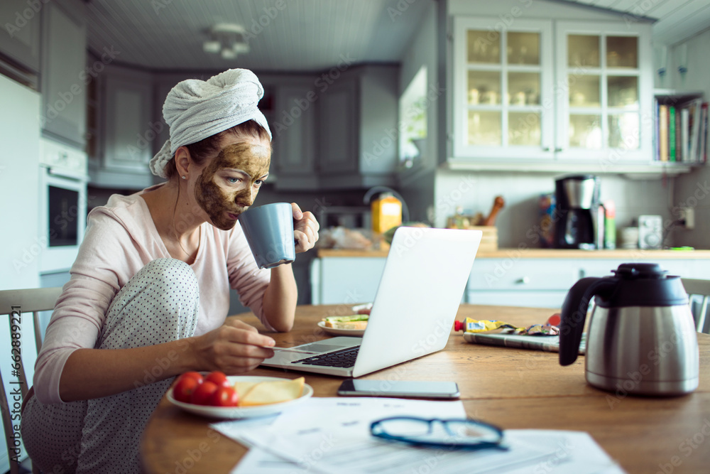Young woman with a cosmetic mask on her face drinking coffee and doing financials on the laptop in the kitchen at home Young woman with a cosmetic mask on her face drinking coffee and doing financials on the laptop in the kitchen at home