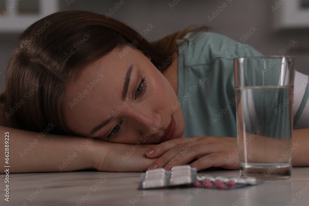 Depressed woman at table with antidepressant pills and glass of water indoors Depressed woman at table with antidepressant pills and glass of water indoors