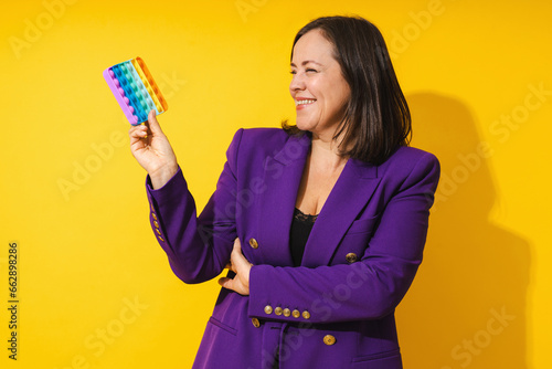 Cheerful middle aged woman wearing purple blazer playing pop-it fidget toy against yellow background
