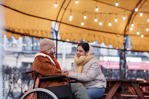 Side view portrait of smiling young woman holding hands with man in wheelchair enjoying date outdoors in winter