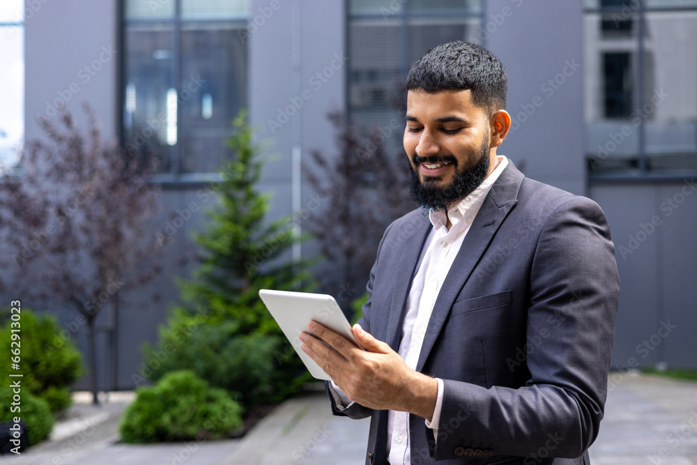 Young smiling Muslim male businessman standing outside office center and using tablet, typing message, chatting, communicating