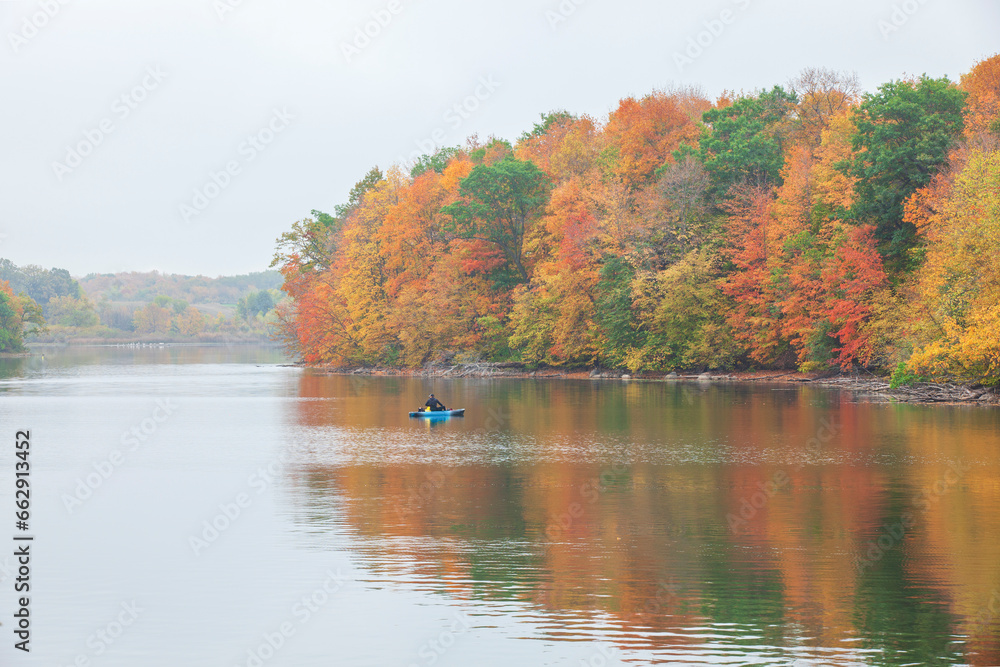 Lone fisherman in a kayak fishes near trees in beautiful autumn color on a calm overcast day Lone fisherman in a kayak fishes near trees in beautiful autumn color on a calm overcast day