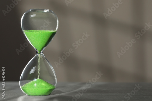 Hourglass with green flowing sand on table against light grey background, space for text