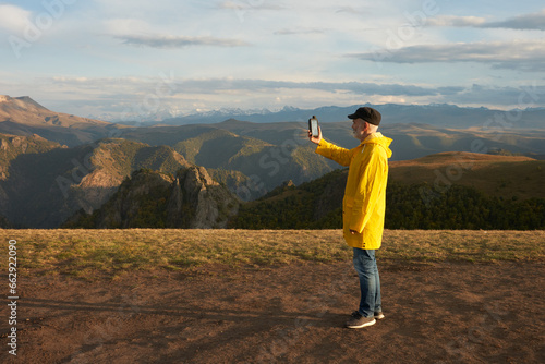 A grown man in a yellow raincoat holding a GPS navigator on a picturesque background of mountainous terrain in the soft light of the setting sun. Copy space