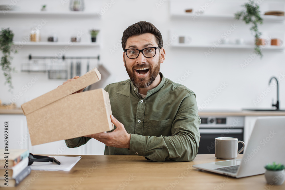 Excited caucasian man in casual wear and glasses sitting at home office with cardboard box in hands. Bearded male blogger happy to receive order from online store during holiday discount.