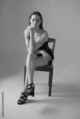 Black and white portrait of a brunette girl without make up on a grey backgroung in a black summer clothes. Woman is sitting on a chair