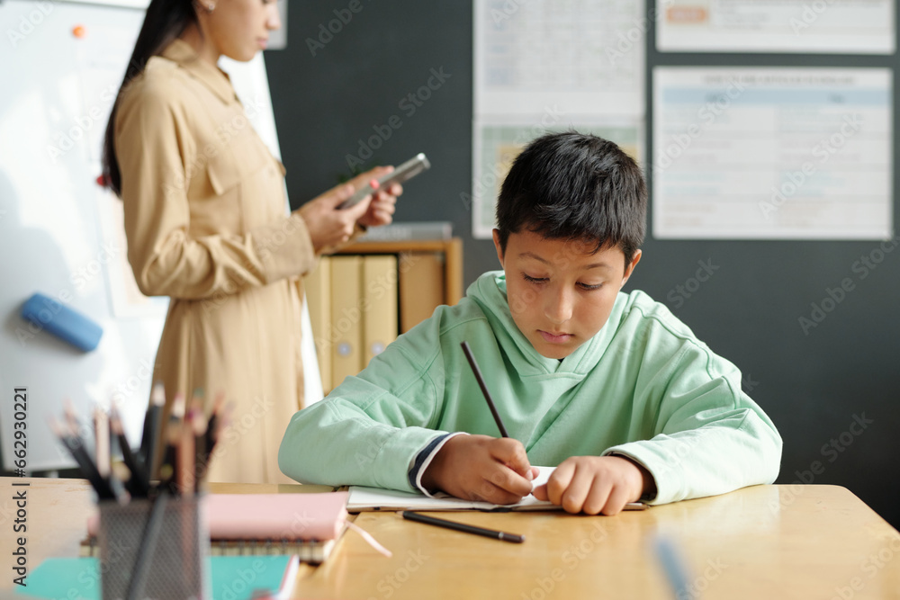 Focus on clever and diligent schoolboy writing down new English grammar rules in copybook while sitting by desk against teacher with tablet