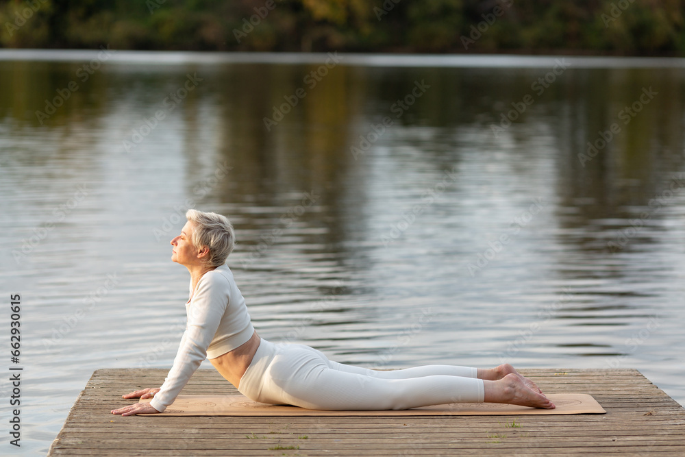 A mature woman with a short haircut in a white suit is doing yoga on the riverbank. Bhujangasana. Upward Dog A mature woman with a short haircut in a white suit is doing yoga on the riverbank. Bhujangasana. Upward Dog