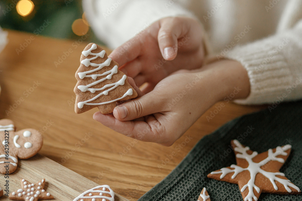 Hands holding gingerbread cookie tree with icing on rustic wooden table. Atmospheric Christmas holiday traditions, family time. Woman decorating cookies with sugar frosting - obrazy, fototapety, plakaty 